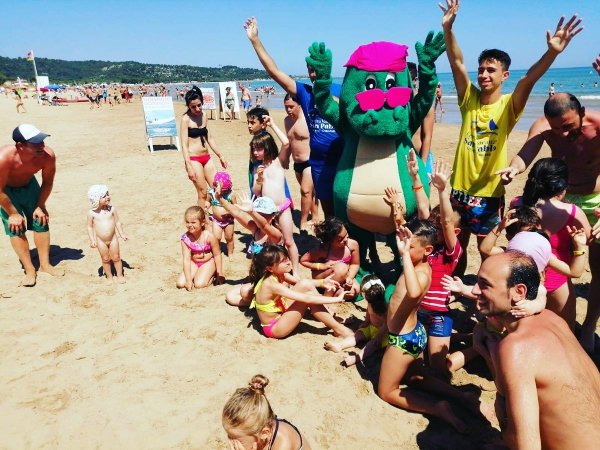 Bambini giocano in spiaggia con una mascotte verde.