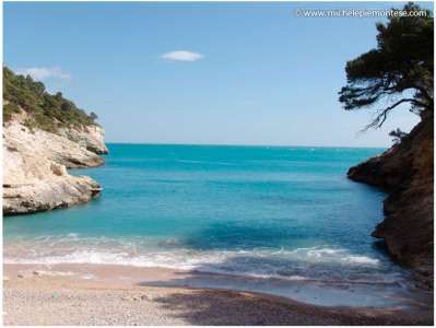 Una baia con acqua turchese, circondata da scogli e alberi, sotto un cielo azzurro.