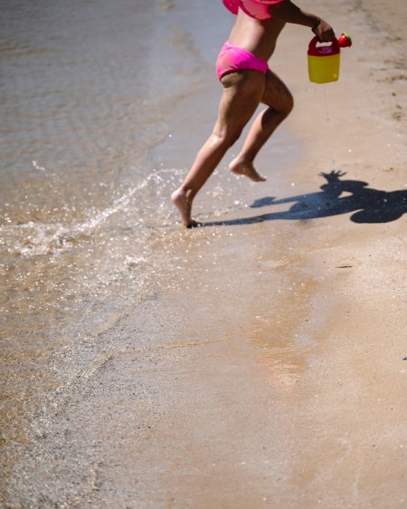 Bambina corre sulla spiaggia con un secchiello giallo.