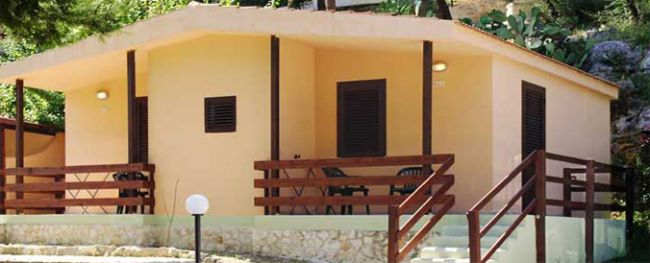 Bungalow con pareti gialle, terrazza in legno, immerso nel verde.