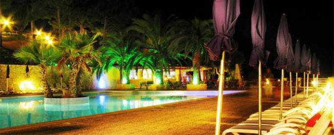 Piscina illuminata di notte, palme illuminate e ombrelloni chiusi.