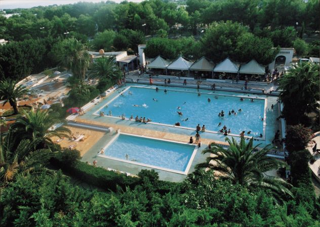 Piscina all'aperto con persone che nuotano, circondata da vegetazione.